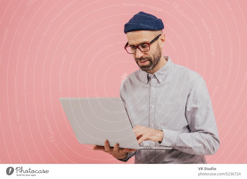 Professional male worker searches for interesting movie to watch, uses gadget, types information on laptop computer, wears formal clothes, isolated over pink background. Online communication