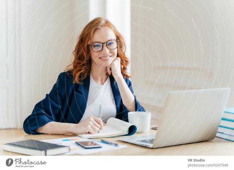 Successful economist in eyewear, checks information on laptop computer, reads information on financial website, writes in notepad and drink hot coffee, sits at workplace. Business and work concept