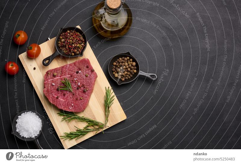 Raw piece of beef with spices pepper, rosemary sprig, salt and olive oil on a wooden board, black background. Copy space raw food cooking preparation gourmet