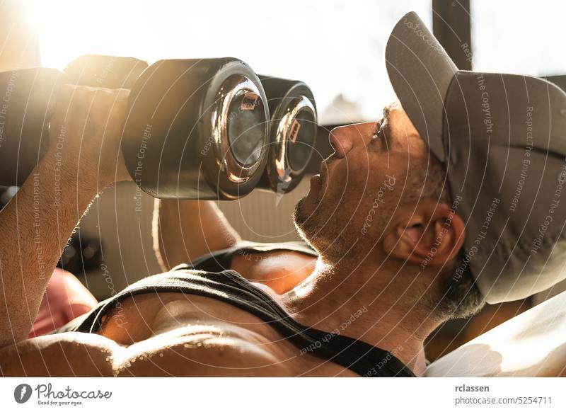 Strong man, bodybuilder exercising with dumbbells in a gym cap sunlight fit workout sport exercise weight studio abdomen lifting power person movement strength