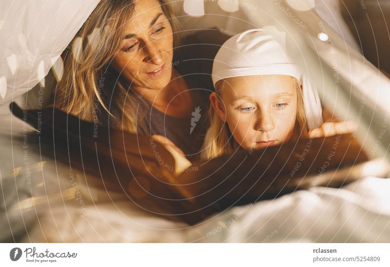 mother and child girl reading a book in bed under blanket before going to sleep. mom kid read book happy child family story flashlight parent concept bedroom
