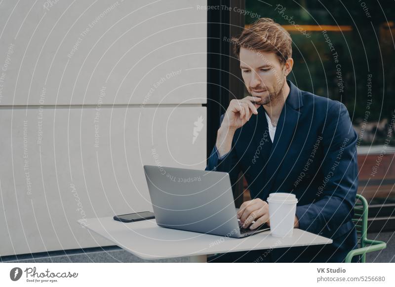 Young man sitting at window and working on laptop. Bearded man sitting at  desktop with smartphone in his hands. Online education. Stock Photo