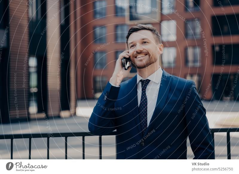 Male office worker enjoying pleasant talk on phone while standing next to office building outdoors handsome businessman talking mobile call cheerful happy good