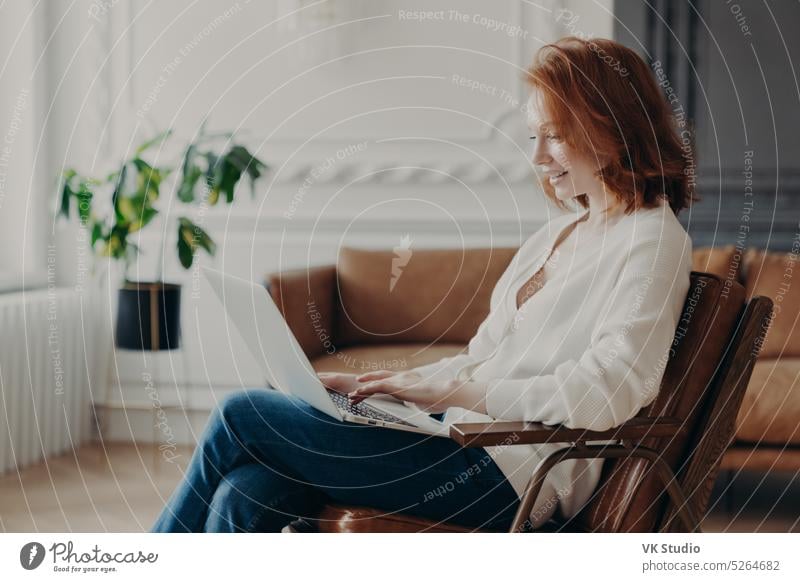 Smiling red haired woman freelancer sits in armchair, keyboards on laptop computer, uploads necessary material for developing site, poses in modern apartment, connected to wireless internet.
