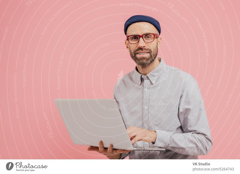 Photo of guy internet user browses website on laptop computer, connected to wireless internet, holds electronic gadget, wears spectacles, formal shirt, isolated over pink background with free space