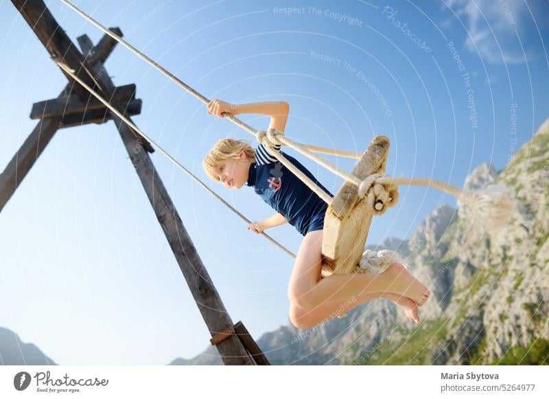 Little boy having fun on large swing on the shore in the Boka Kotor Bay of the Adriatic Sea in the Balkan Mountains, Montenegro. Happy child on a sea resort on sunny summer holidays day.