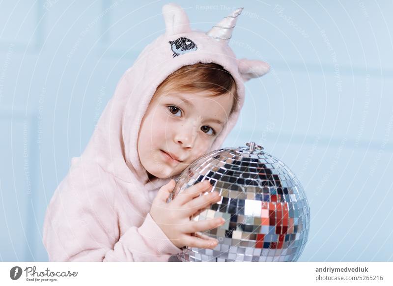 Smiling beautiful little girl in unicorn pink costume is smiling and having fun with disco ball on a blue wall background. happy childhood. hat face cute kid
