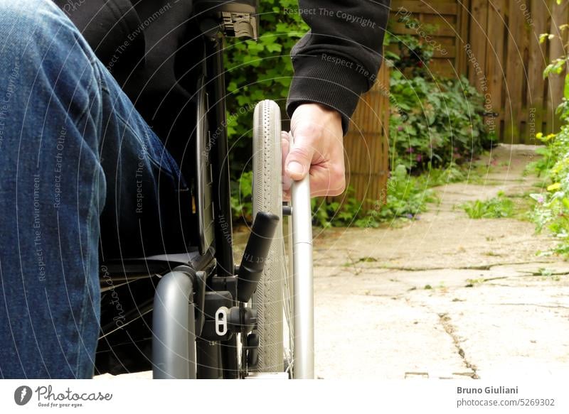 A disabled person in a wheelchair. A man with reduced mobility in an alley with vegetation. alone disabilities disability outdoors hand close up