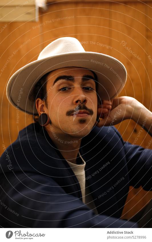 Stylish ethnic guy looking at camera near wooden wall man style model cool personality confident portrait male young hispanic hat dark hair mustache earplug