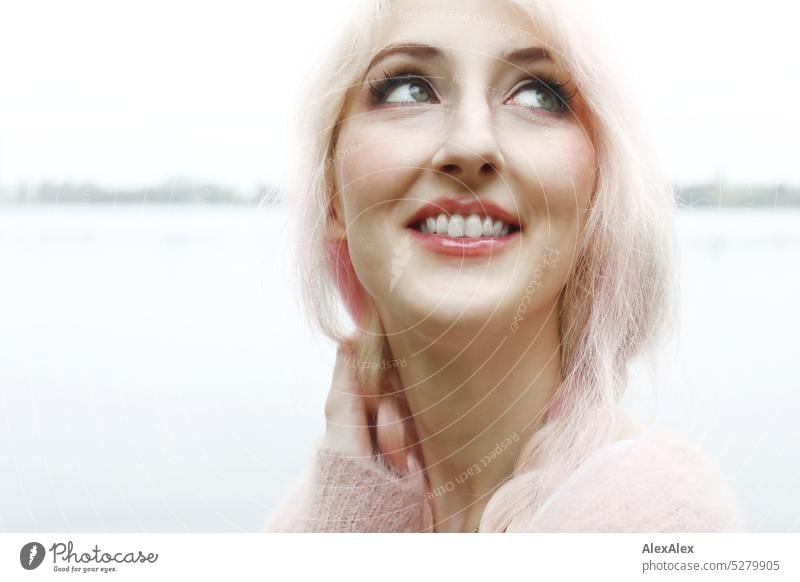 Young woman with pink blonde hair looks at sky and smiles in front of lake with lakeside in distance Woman portrait pretty Slim long hairs pink hair Pink Blonde