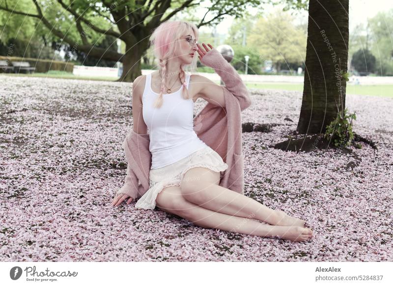 Young woman with pink blonde hair sits barefoot on the ground of park covered with pink cherry blossom petals Woman portrait pretty Slim long hairs pink hair