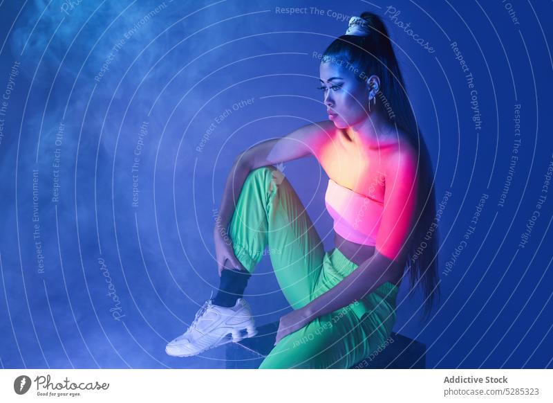 Fit mixed race woman sitting on cube in smoke model projector posture studio shot appearance neon illuminate lady young ethnic female outfit gorgeous sporty