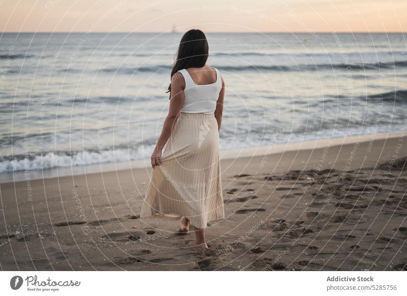 Anonymous young ethnic woman standing on sandy seashore during summer vacation walk beach enjoy happy holiday sunset traveler nature female barefoot long hair