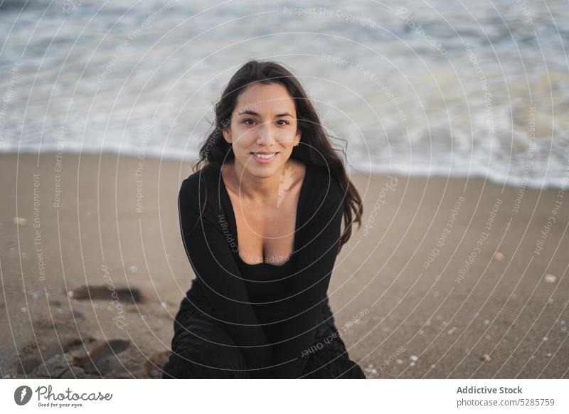 Delighted young ethnic female tourist sitting on haunches on beach and smiling woman self assured vacation holiday feminine sea rest smile touch cheek portrait