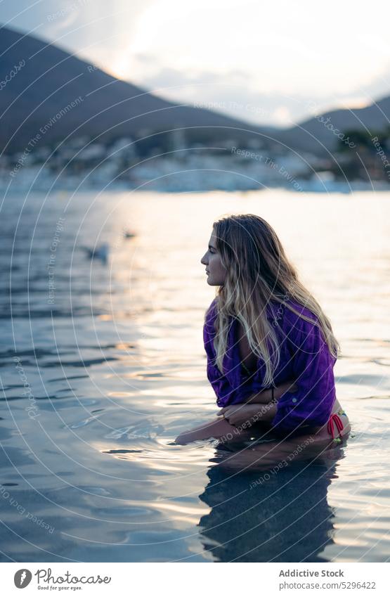 Calm young woman sitting in sea water and looking away beach sunset dreamy vacation holiday recreation admire summer seashore nature relax female long hair