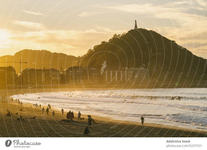 Tourists walking on seashore in sunset people beach resort mountain evening building wave coast sundown water dusk nature ocean sand vacation stroll summer sky