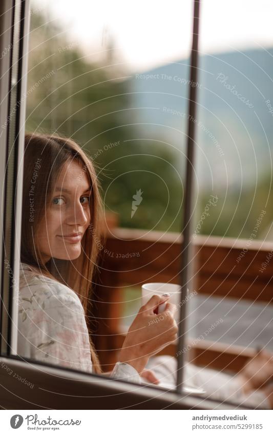 Young female standing after taking a shower in the morning on balcony of the hotel. holding a cup of coffee or tea in her hands. Looking outside nature forest and Mountain