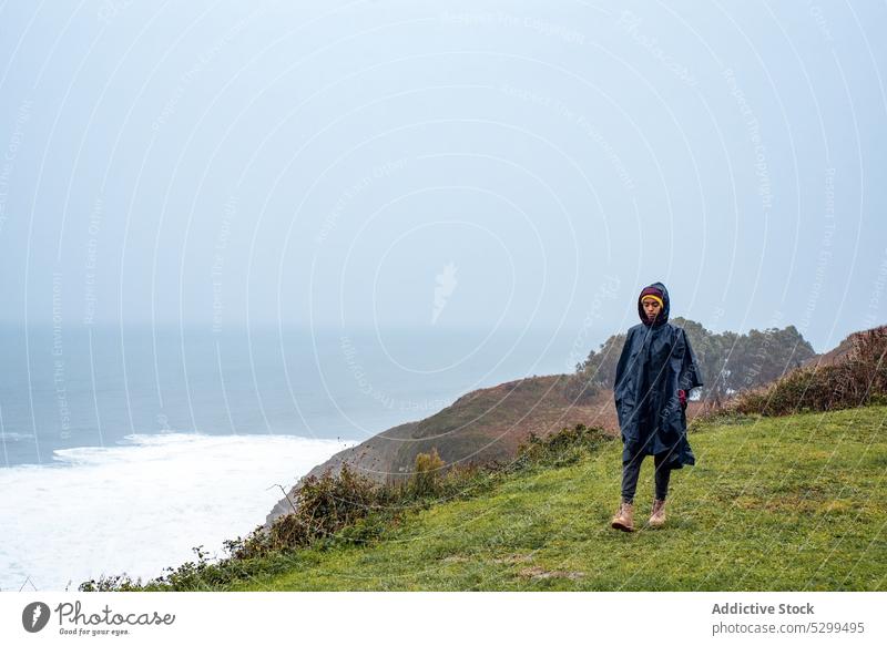 Woman in raincoat walking on mountain near ocean woman tourist traveler hike cliff sea vacation trip female coast weekend shore grass nature journey blue sky