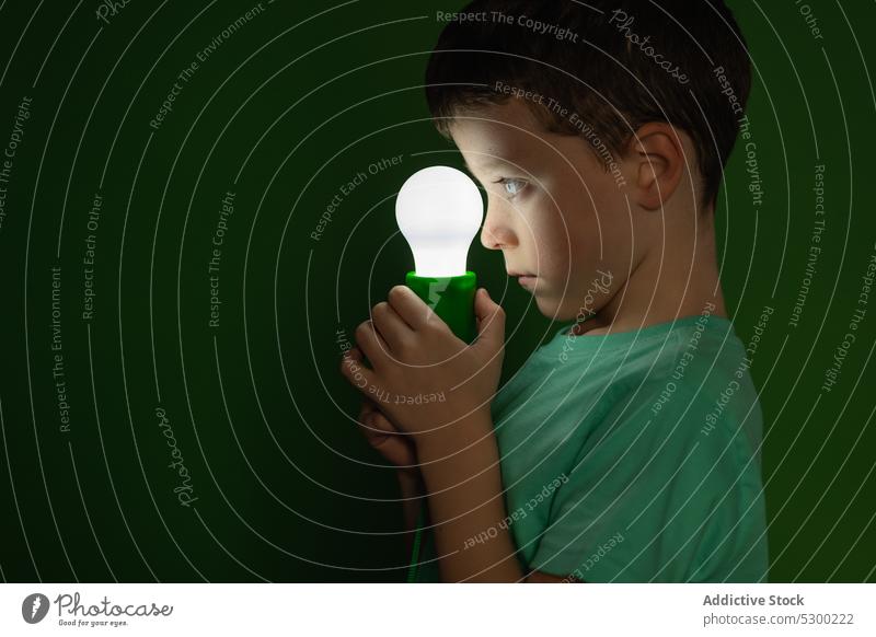 Little boy with light bulb in dark room child kid curious lamp glow illuminate studio shot cute adorable preteen childhood close stand little green happy home