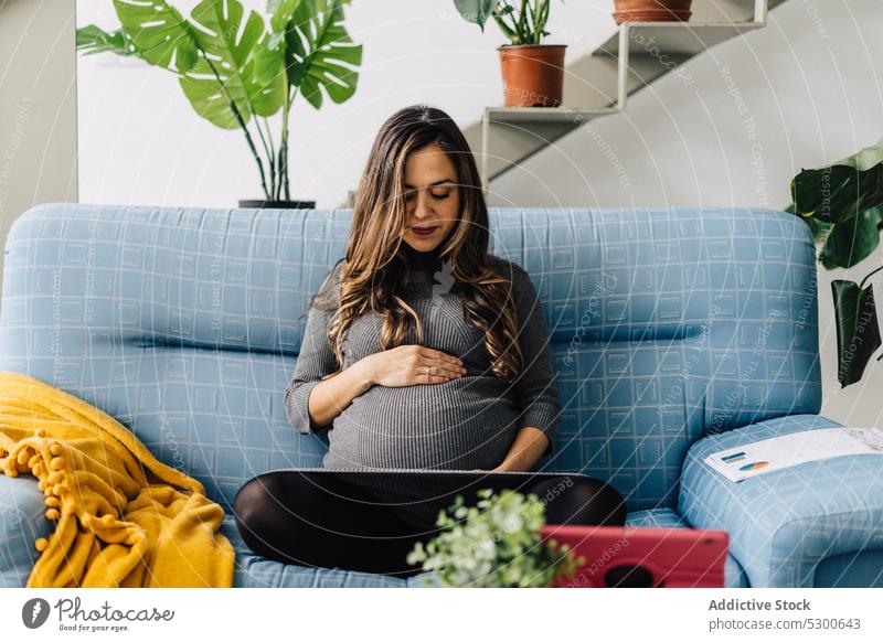Smiling pregnant woman watching film on tablet movie maternity leave leisure free time sofa at home smile female young connection mother checkered wireless