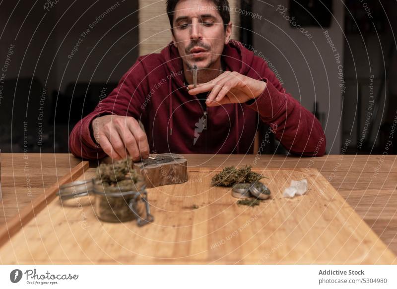 Adult man smoking cannabis at home smoke smoker marijuana light joint blunt match ganja habit male adult medical legalize nicotine weed drug wooden table addict