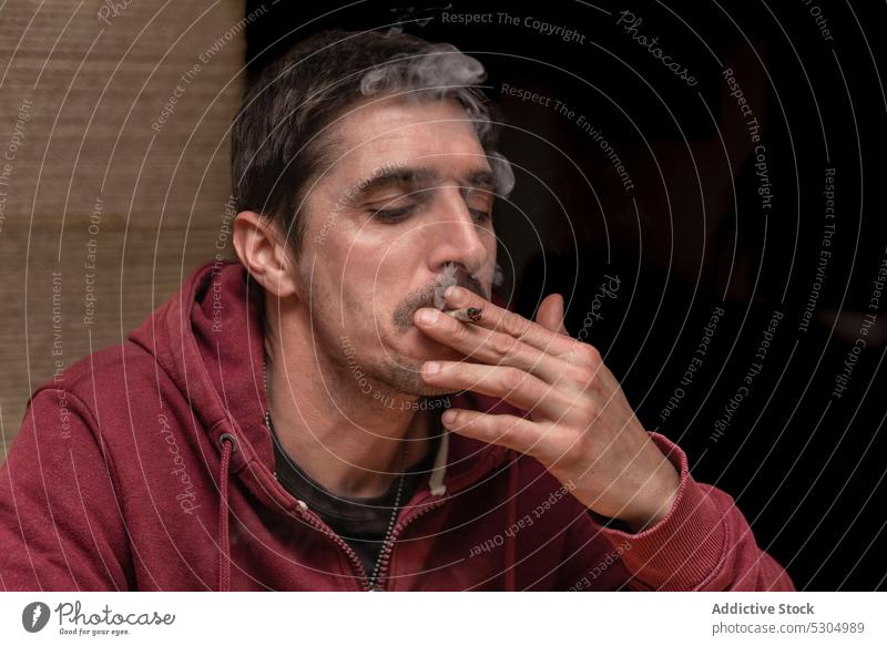 Adult man with mustache smoking marijuana blunt smoke smoker inhale medical joint light drug cannabis male adult narcotic hemp habit at home hoodie addict