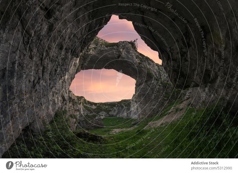 Double hole cave in mountainous terrain against sunset sky rocky formation green grass colorful picturesque sundown landscape scenery vivid vibrant breathtaking