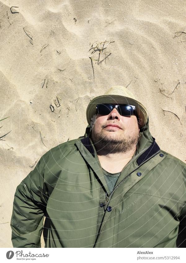 bearded man with straw hat, sunglasses and green weather jacket is lying in the dune sand on a sunny day by the sea Man Holidaymakers hikers Summer spring