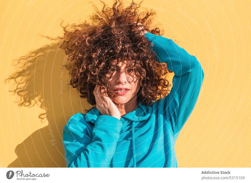 Charming curly haired woman touching cheek against yellow wall calm portrait style individuality color touch cheek touch hair charming outfit female appearance