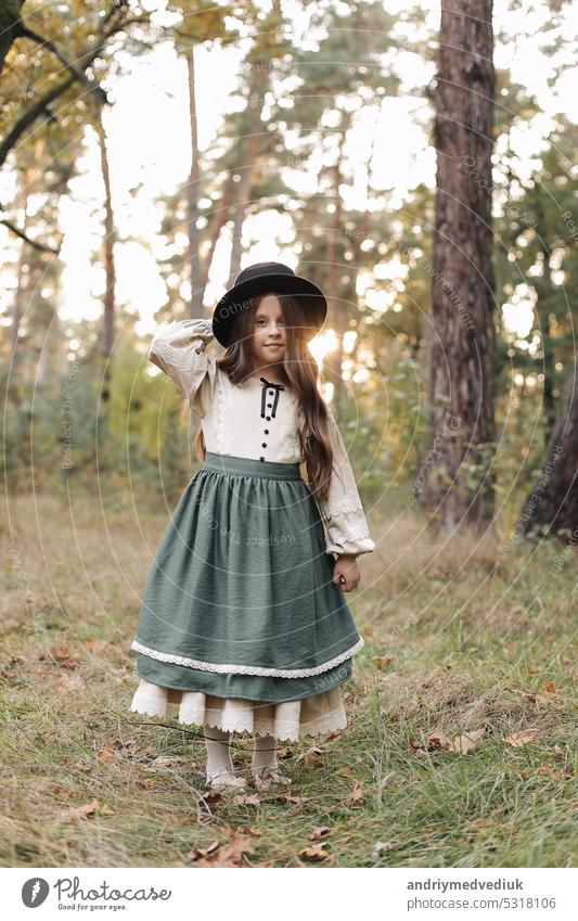 Portrait of little caucasian girl looking at camera standing outdoors. Stylish adorable smiling child in black hat enjoying childhood testimonial concept at green park background.