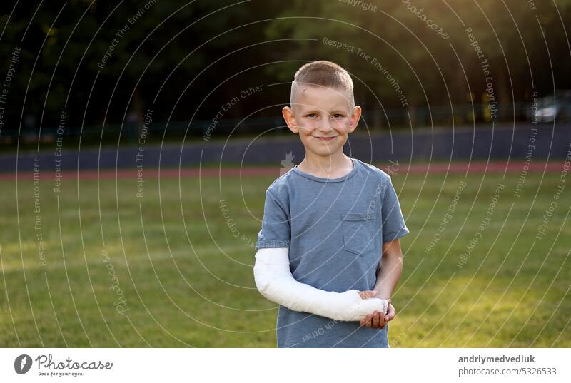 Smiling little boy with broken hand looks at camera outdoors on sports ground on summer day. 9 years child holding his healthy hand another fractured limb with plaster cast on the arm. Health concept