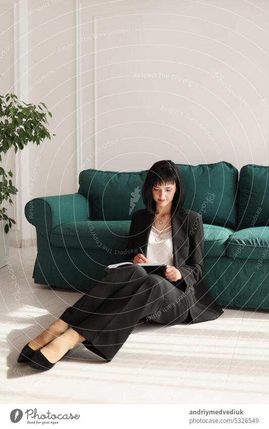 smiling businesswoman writes in note pad and looking in camera. brunette woman is sitting close to green sofa in a black suit and high heels shoes. Green houseplant in interior. Work from home.