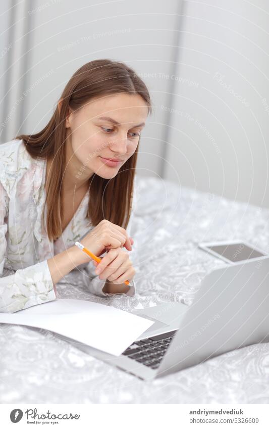 Beautiful young woman working, using laptop computer and makes notes in the bedroom at home. Freelancer. Writing, typing. Girl checking social apps. Communication and technology concept