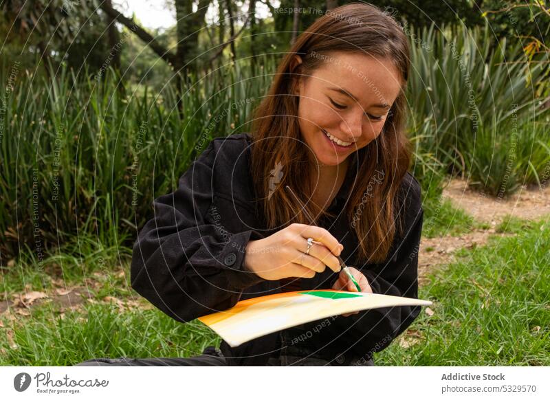 Cheerful young woman drawing in nature paint artist painter cheerful rest happy smile hobby inspiration paintbrush creative paper sit plant talent casual grass