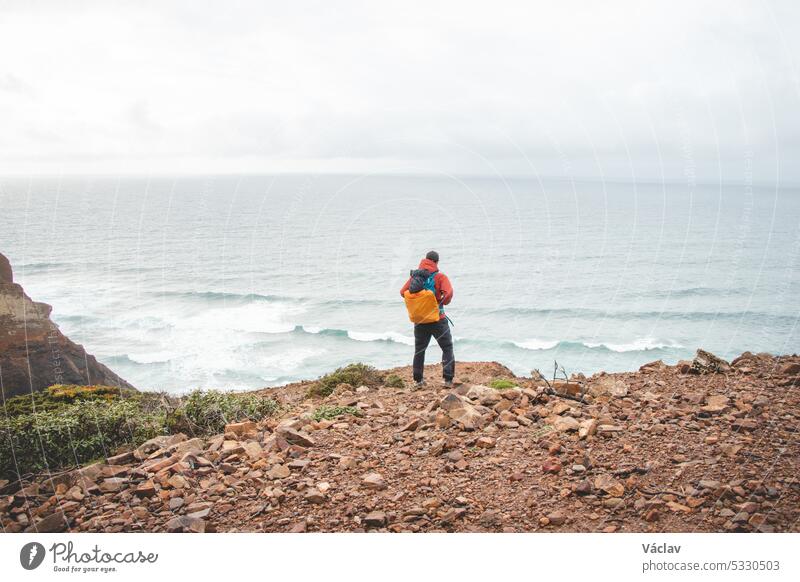 Adventurous man standing on the edge of a cliff enjoys the view of the Atlantic coast in the Odemira region of southwestern Portugal. Wandering the Rota Vicentina