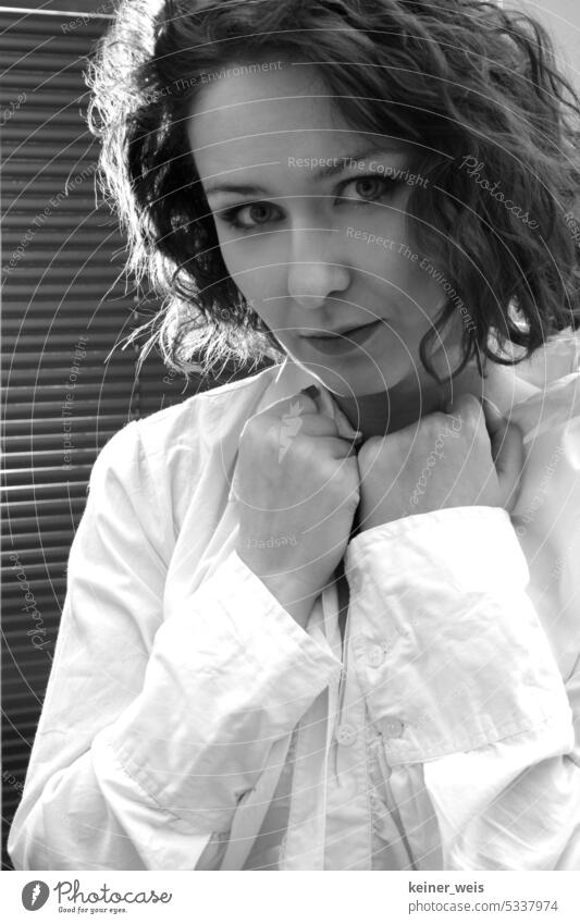 Woman with curly hair stands in white shirt with hands on shirt collar in front of blinds on window as portrait in black and white Hair curls Shirt White
