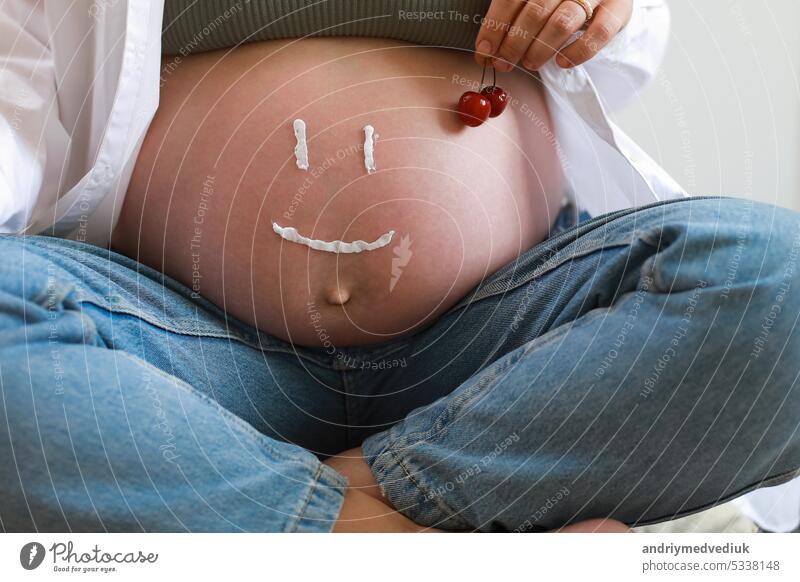 Close-up of torso of young pregnant model applying moisturizer on her belly to avoid stretch marks. Future mom with funny smile from moisturizing cream on her tummy. Pregnancy skincare concept