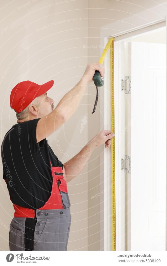 Worker in red cap and work suit with the measuring tape. Man is installing the doors. Measure tape in hands. Repair works. Maintenance in the apartment. man