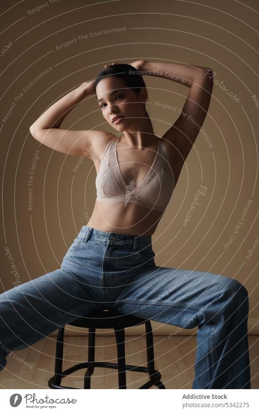 Graceful woman sitting on chair in light studio style lingerie portrait model thoughtful sensual stool jeans fashion underwear figure arms raised female