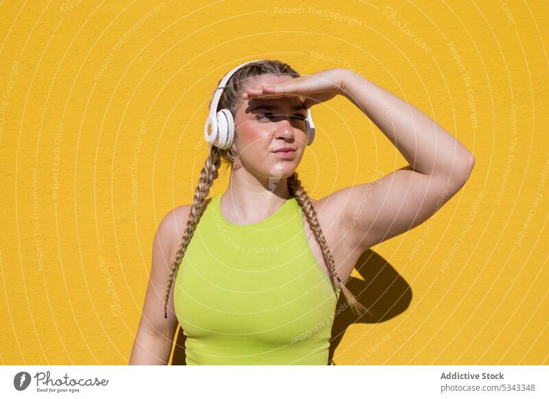 Young woman standing with headphones near yellow backdrop listen music sunlight audio using focus wireless summer melody song bright gadget device female young