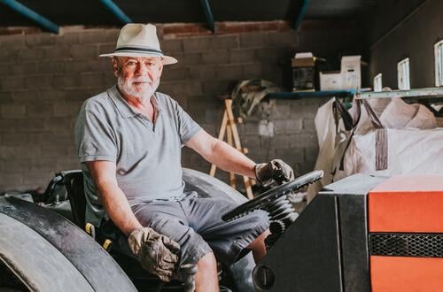 Aged male farmer sitting on tractor in garage man portrait senior countryside agriculture machinery aged rural vehicle shabby glove hat sunhat gray beard