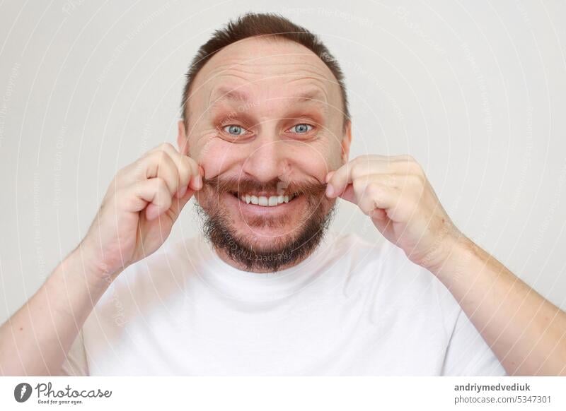 Portrait of happy caucasian bearded man touches his moustache in white casual t shirt smiles and looks at camera on white background. Lifestyle and positive emotions concept. Kind and happy face