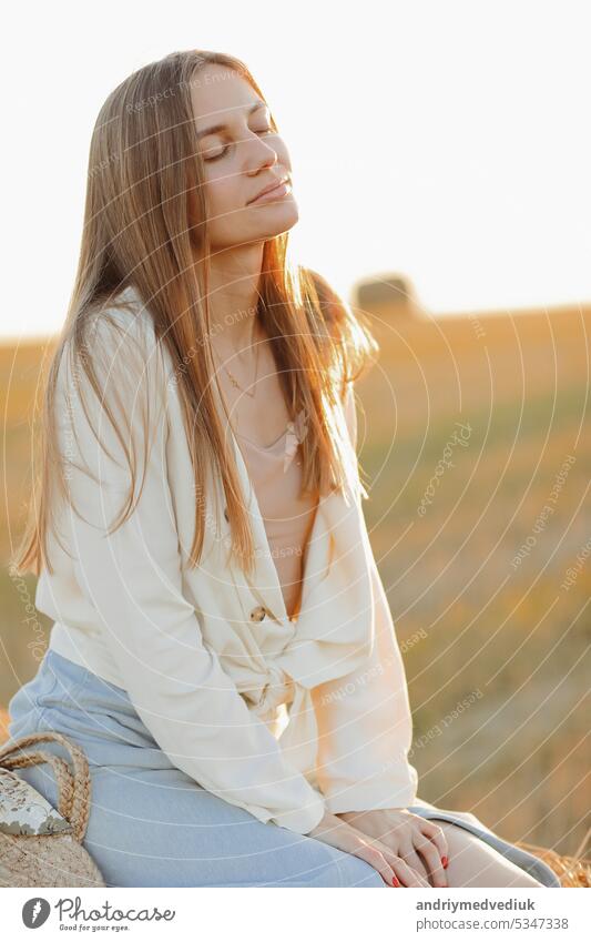 Beautiful portrait of a young woman during the sunset with warm yellow sun rays on her face with bales of straw on the background rural beautiful countryside