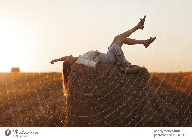 Young woman with long hair, wearing jeans skirt, light shirt is lying on straw bale in field in summer on sunset. Female portrait in natural rural scene. Environmental eco tourism concept.