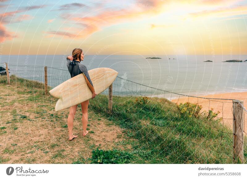 Surfer woman putting her hair up while looking at the beach from the shore surfer young surfboard surf board sunset sunlight copy space wetsuit coast people