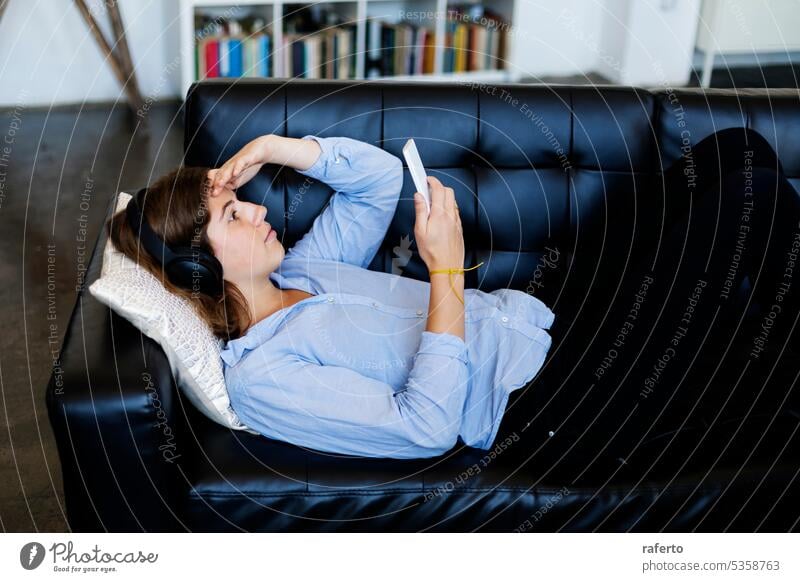 A Young woman stretched out on leather sofa with headphones, using mobile phone young woman technology relaxation leisure indoor home comfort trendy stylish