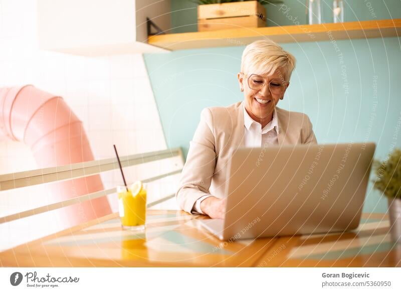 Senior woman using mobile phone while working on laptop and drinking fresh orange juice in the cafe aged call coffee coffee shop coffeehouse communication