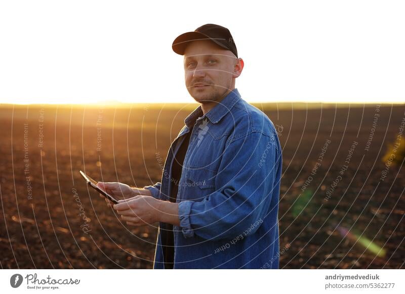 Smiling male farmer holds digital tablet on field with plowed soil at sunset and looks at camera. Checking and control of soil quality, land readiness for sowing crops. Smart farming technology.