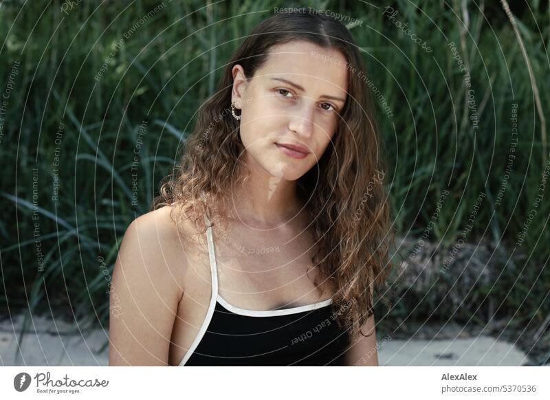 direct portrait of young beautiful brunette smiling woman sitting in the sand in front of a forest warm Summery Beautiful weather plants proximity Day