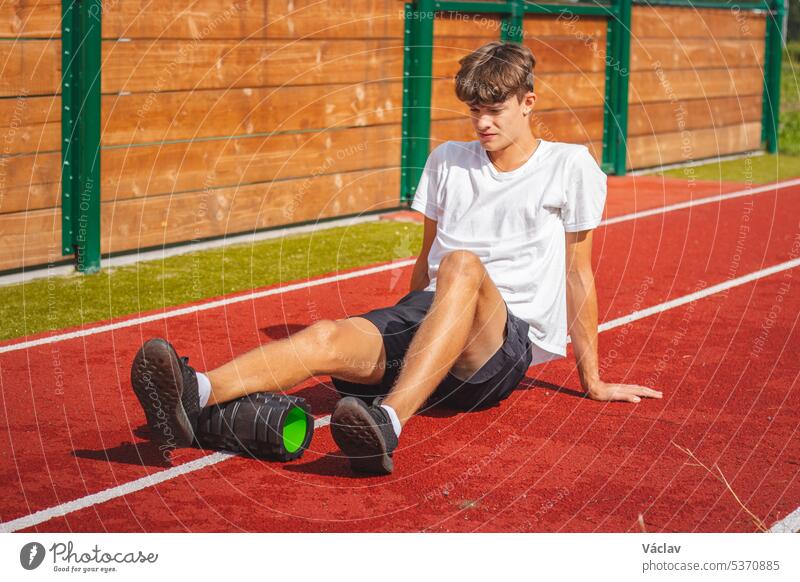 Brown-haired boy with an athletic physique on an athletic oval massages his muscles with a foam roller for better recovery. Post-workout strength abdominal